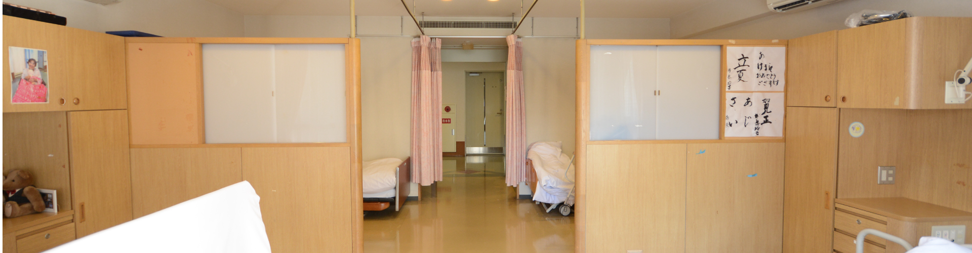 木製パネル仕切りの多床室。奥に浴室への通路が見える介護施設の居室